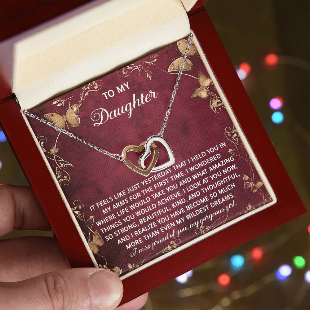 A hand holds an open jewelry box with the Daughter-Wildest Dreams - Interlocking Hearts Necklace. The heart-shaped pendant is adorned with dazzling cubic zirconia crystals. Inside, a loving message for a daughter on a decorative background makes it an ideal keepsake.