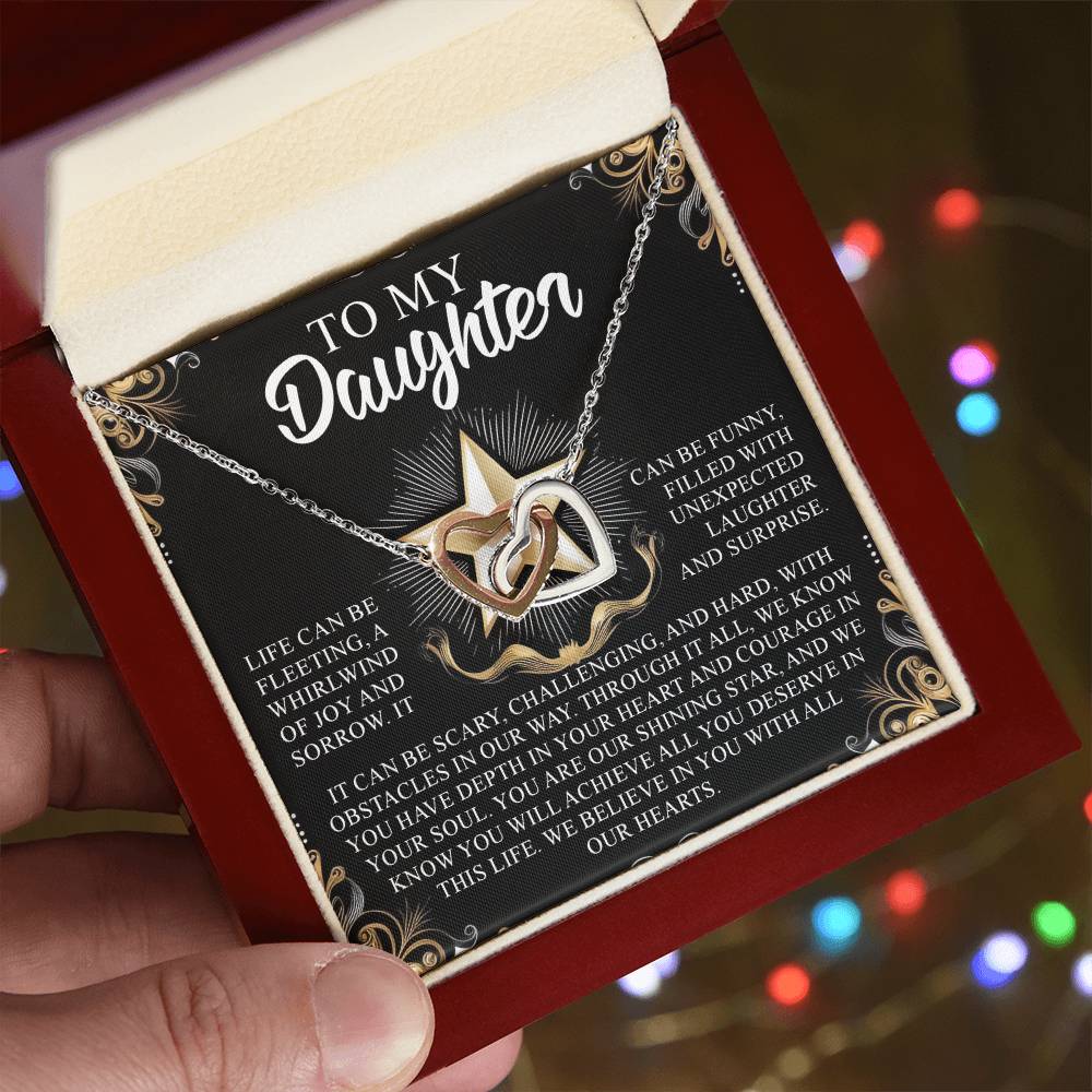 An open gift box displays the "Daughter-Shining Star - Interlocking Hearts Necklace" with cubic zirconia. The card says "To My Daughter," sharing a heartfelt message about life's challenges and support, amidst festive, blurred lights in the background.