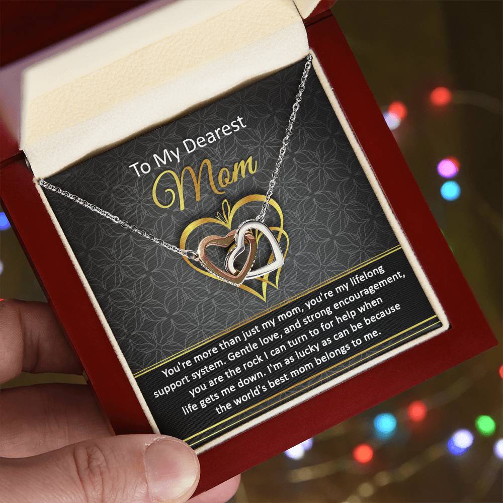 Close-up of the "To Mom, Belongs To Me2 - Interlocking Hearts Necklace" with cubic zirconia crystals in a gift box, featuring a heartfelt message to "Mom." Blurred colorful lights in the background enhance this personalized gift.