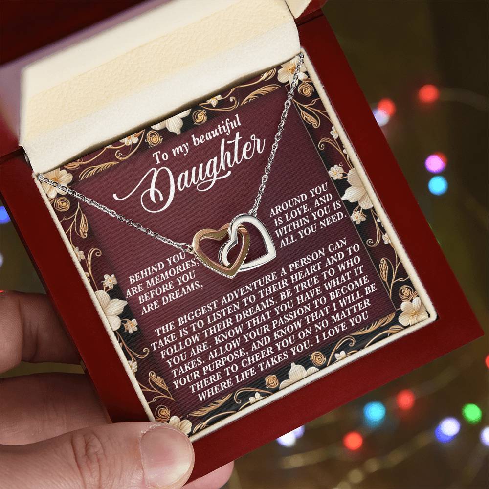 An open gift box reveals the "Daughter-Follow Dreams - Interlocking Hearts Necklace" with cubic zirconia crystals. The lid displays a floral border and a loving message to a daughter about pursuing dreams, against a backdrop of blurred festive lights.