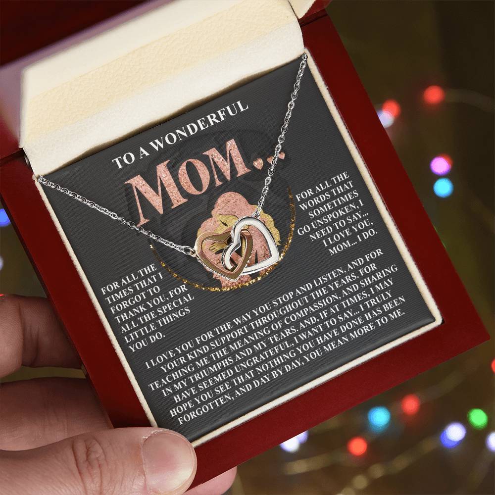 A hand holds an open gift box with a Mom-More To Me 2 - Interlocking Hearts Necklace adorned with cubic zirconia, with the lid featuring a sentimental message for Mom. Blurred colorful lights in the background make it perfect for an anniversary gift.