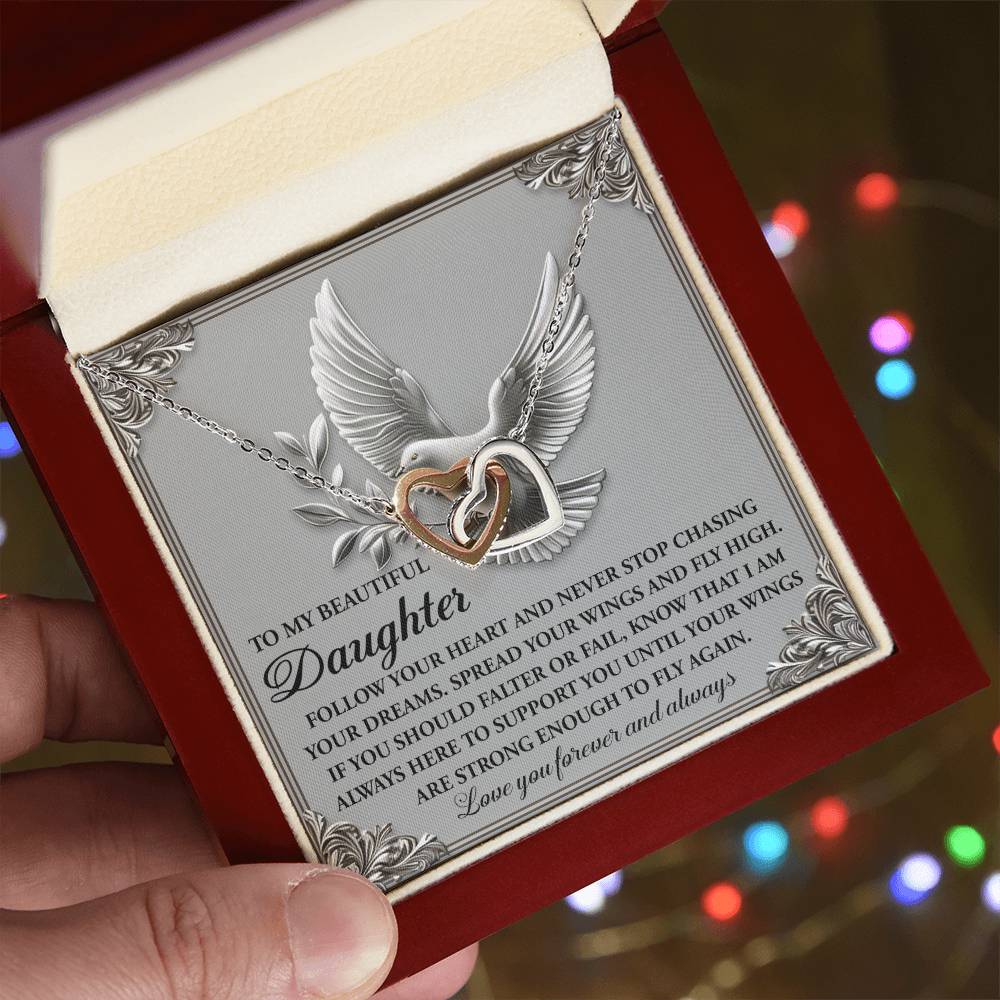 A hand unveils an open jewelry box containing the Daughter-Your Wings - Interlocking Hearts Necklace, featuring a dove-accented heart and infinity pendant. An ornate message to a daughter is inscribed, with cubic zirconia crystals glimmering as colorful lights blur in the background.