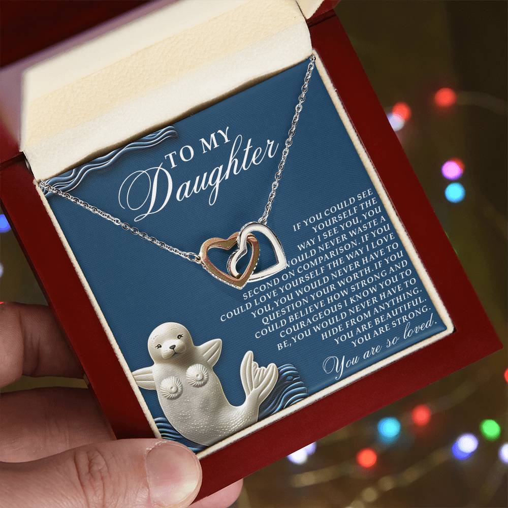 A hand holds an open jewelry box displaying the Daughter-So Loved - Interlocking Hearts Necklace in rose gold with cubic zirconia, accompanied by a heartfelt message to a daughter and a seal image, set against blurred colorful lights.