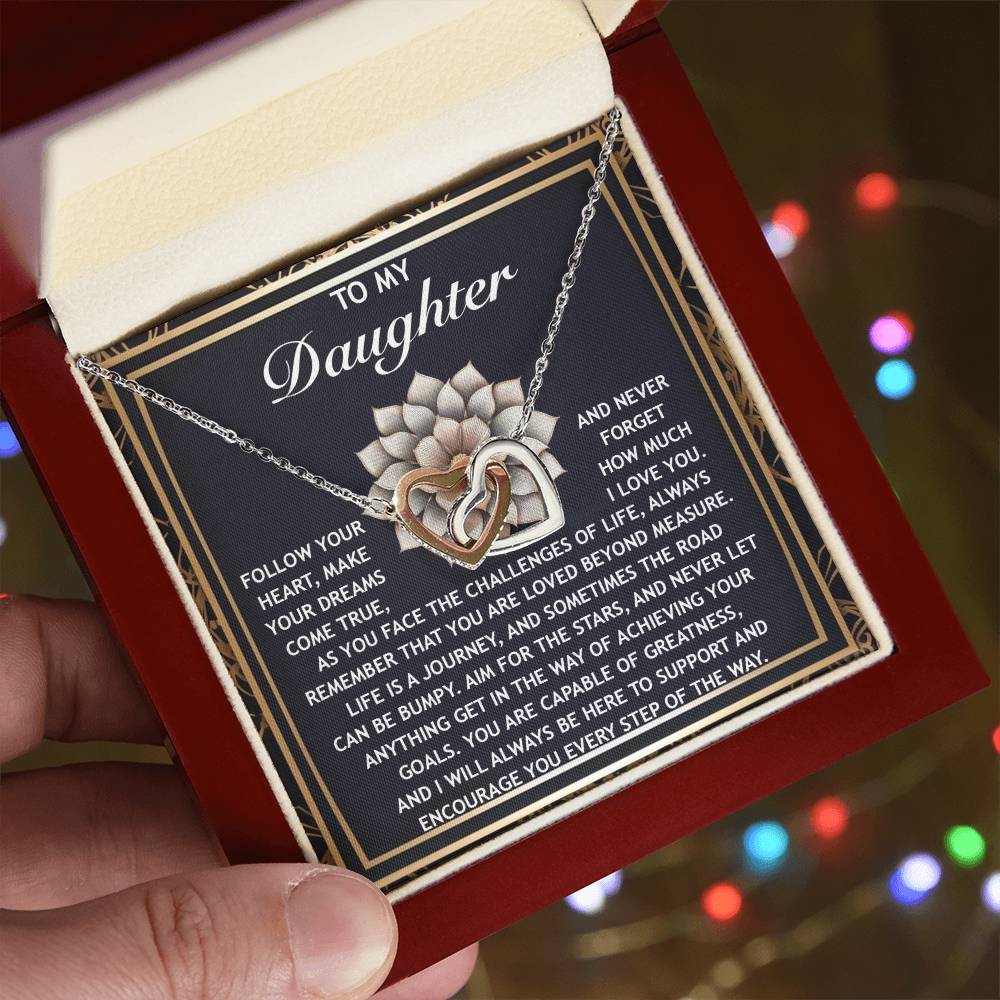 An open jewelry box showcases the "Daughter-In The Way - Interlocking Hearts Necklace" with a heart-shaped pendant, featuring an inspirational message, elegantly rose gold finished and sparkling with cubic zirconia crystals against a blurred colorful background.
