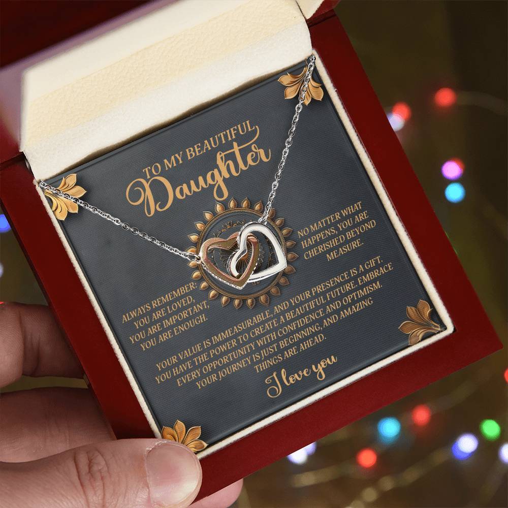 An open jewelry box reveals the "Daughter-Beautiful Future - Interlocking Hearts Necklace" featuring a silver finish. The intertwined hearts, adorned with cubic zirconia crystals, are engraved with supportive words for a daughter and framed by subtle decorative elements.