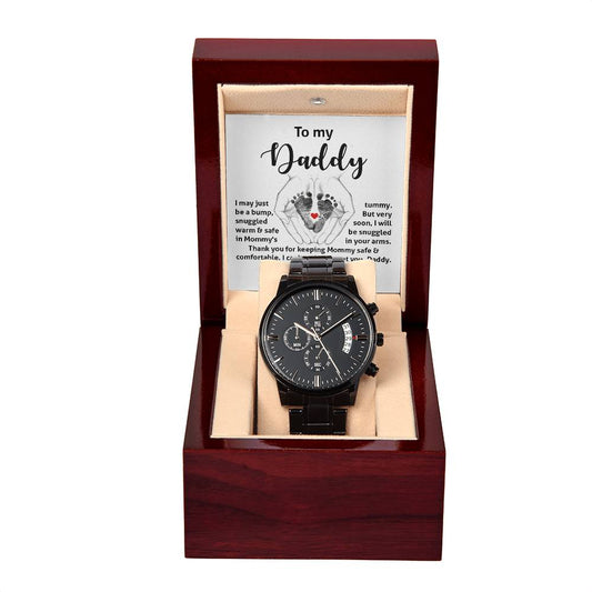 A black chronograph wristwatch with a round dial displayed in an open red wooden box, featuring a sentimental message to "daddy" on the inner lid.