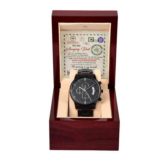 A black chronograph watch with a metal band is presented in an open wooden box with a beige interior. Behind the watch, a card with a note is seen.