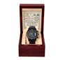 A black chronograph watch with a metal band is presented in an open wooden box with a beige interior. Behind the watch, a card with a note is seen.