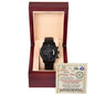 A black chronograph watch with a metal band is presented in an open wooden box with a beige interior. Behind the watch, a card with a note is seen.