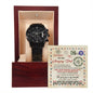 A black chronograph watch with a metal band is presented in an open wooden box with a beige interior. Behind the watch, a card with a note is seen.