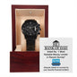 A "To Dad, Financial Burden - Metal Chronograph Watch" with a day and date display is placed inside an open wooden watch box. The inside cover of the box has a humorous message that reads, "Bank of Dad: Voted No. 1 Most Reliable Money Lender.