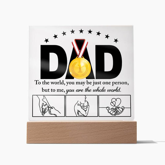 A framed acrylic plaque with the word "DAD" and a gold medal sits elegantly on an LED wooden base. Text underneath reads, "To the world, you may be just one person, but to me, you are the whole world," above three hand-drawn images of hands. A perfect sentimental gift for any father. This is the To Dad, The Whole World - Acrylic Square Plaque.