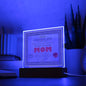 The "To Mom, World's Greatest Mom" Acrylic Square Plaque, illuminated on a dimly lit LED wooden base, shines brightly amid books and a plant in soft blue lighting.