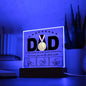 A framed acrylic plaque with the word "DAD" and a gold medal sits elegantly on an LED wooden base. Text underneath reads, "To the world, you may be just one person, but to me, you are the whole world," above three hand-drawn images of hands. A perfect sentimental gift for any father. This is the To Dad, The Whole World - Acrylic Square Plaque.