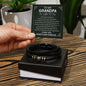 A hand holds a card that reads "To My Grandpa" above a decorative message. Below the card is a black vegan leather bracelet with metal accents in a white box—an ideal engraved gift for any special man, the "To Grandpa, Thank You - Love You Forever Bracelet.