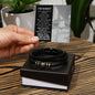 A hand holding an apology card over an open black box containing the "Sorry-Wish I Could - Love You Forever Bracelet," a black braided men's bracelet with metallic accents. The card features a heartfelt apology message, making this engraved bracelet a thoughtful gesture for any occasion.