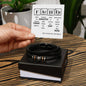 A hand holding a periodic table-inspired card with a humorous "FATHER" message. Below the card, an engraved men's bracelet made of black braided leather with metallic accents, labeled "To Father, Still The Best - Love You Forever Bracelet," is displayed in an open gift box.