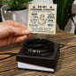 A person holding a note that reads "To My Awesome Husband" over a black braided vegan leather bracelet with metallic beads and symbols, placed in an open gift box labeled "To My Husband, Battle For You - Love You Forever Bracelet".