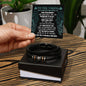 A hand holding a card with an inspirational message addressed "To Dad, Never Giving Up - Love You Forever Bracelet" above a black box containing a braided vegan leather bracelet with metal accents.