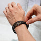 A hand holds a card that reads "To My Grandpa" above a decorative message. Below the card is a black vegan leather bracelet with metal accents in a white box—an ideal engraved gift for any special man, the "To Grandpa, Thank You - Love You Forever Bracelet.