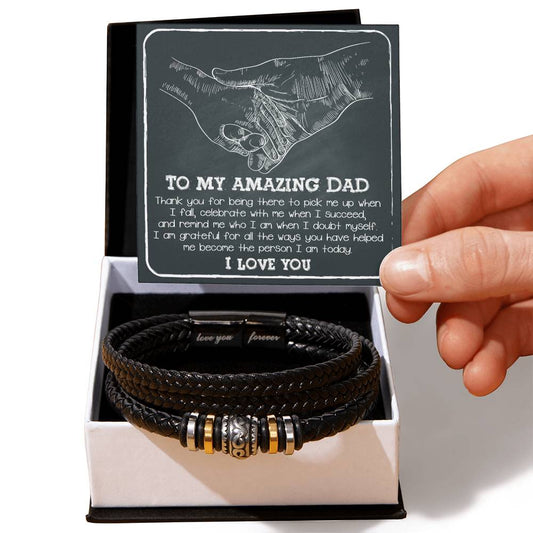 A hand presents an open gift box containing the "To Dad, Pick Me Up" bracelet with metal details, alongside a heartfelt greeting card for a father.