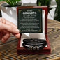 A hand holds a card that reads "To My Grandpa" above a decorative message. Below the card is a black vegan leather bracelet with metal accents in a white box—an ideal engraved gift for any special man, the "To Grandpa, Thank You - Love You Forever Bracelet.