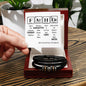 A hand holding a periodic table-inspired card with a humorous "FATHER" message. Below the card, an engraved men's bracelet made of black braided leather with metallic accents, labeled "To Father, Still The Best - Love You Forever Bracelet," is displayed in an open gift box.
