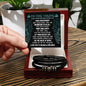 A hand holding a card with an inspirational message addressed "To Dad, Never Giving Up - Love You Forever Bracelet" above a black box containing a braided vegan leather bracelet with metal accents.