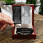 A hand holding an apology card over an open black box containing the "Sorry-Wish I Could - Love You Forever Bracelet," a black braided men's bracelet with metallic accents. The card features a heartfelt apology message, making this engraved bracelet a thoughtful gesture for any occasion.