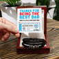 A hand holds a card reading "Thanks for being the best dad" over a wooden box with the To Dad, Gift Voucher - Love You Forever Bracelet, a sleek men's accessory in black leather with stainless steel accents.