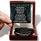 A hand holding a card with an inspirational message addressed "To Dad, Never Giving Up - Love You Forever Bracelet" above a black box containing a braided vegan leather bracelet with metal accents.