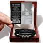 A hand holding an apology card over an open black box containing the "Sorry-Wish I Could - Love You Forever Bracelet," a black braided men's bracelet with metallic accents. The card features a heartfelt apology message, making this engraved bracelet a thoughtful gesture for any occasion.