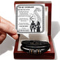 A hand holds a card reading "To My Husband" with a heartfelt message. Below the card, a stylish men's bracelet made of vegan leather with metallic accents and an engraved message rests in a box: the "To My Husband, I Am Yours - Love You Forever Bracelet.