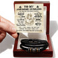A person holding a note that reads "To My Awesome Husband" over a black braided vegan leather bracelet with metallic beads and symbols, placed in an open gift box labeled "To My Husband, Battle For You - Love You Forever Bracelet".