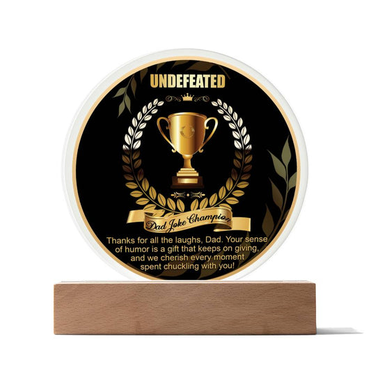 A "To Dad, Dad Joke Champion - Acrylic Circle Plaque" with "UNDEFEATED" at the top, featuring a gold trophy illustration and a banner labeled "Dad Joke Champion." The printed design includes a message about appreciation for dad's humor and is mounted on a wooden base.