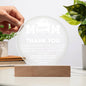 A hand touches the "To Mom, Standing By Me" acrylic circle plaque on a wooden stand with an engraved "MOM THANK YOU" message. This elegant gift suits special occasions and complements home decor, enhanced by two potted plants in the serene background.