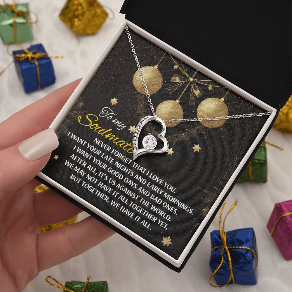 A hand holds an open box containing the Soulmate-Have It All - Forever Love Necklace, featuring a heart-shaped pendant adorned with cubic zirconia set in a 14k white gold finish. The lid of the box showcases a sentimental message and is decorated with a background of golden ornaments.