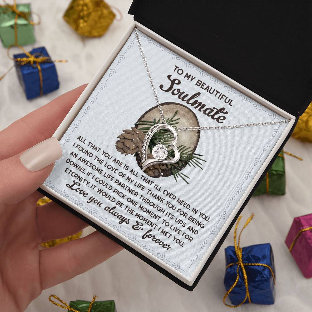 A hand presents an open box revealing the Soulmate-One Moment - Forever Love Necklace, featuring a stunning gold finish, accompanied by a card that reads, "To my beautiful soulmate." Around the box are small wrapped gifts that enhance the sentiment of everlasting love.