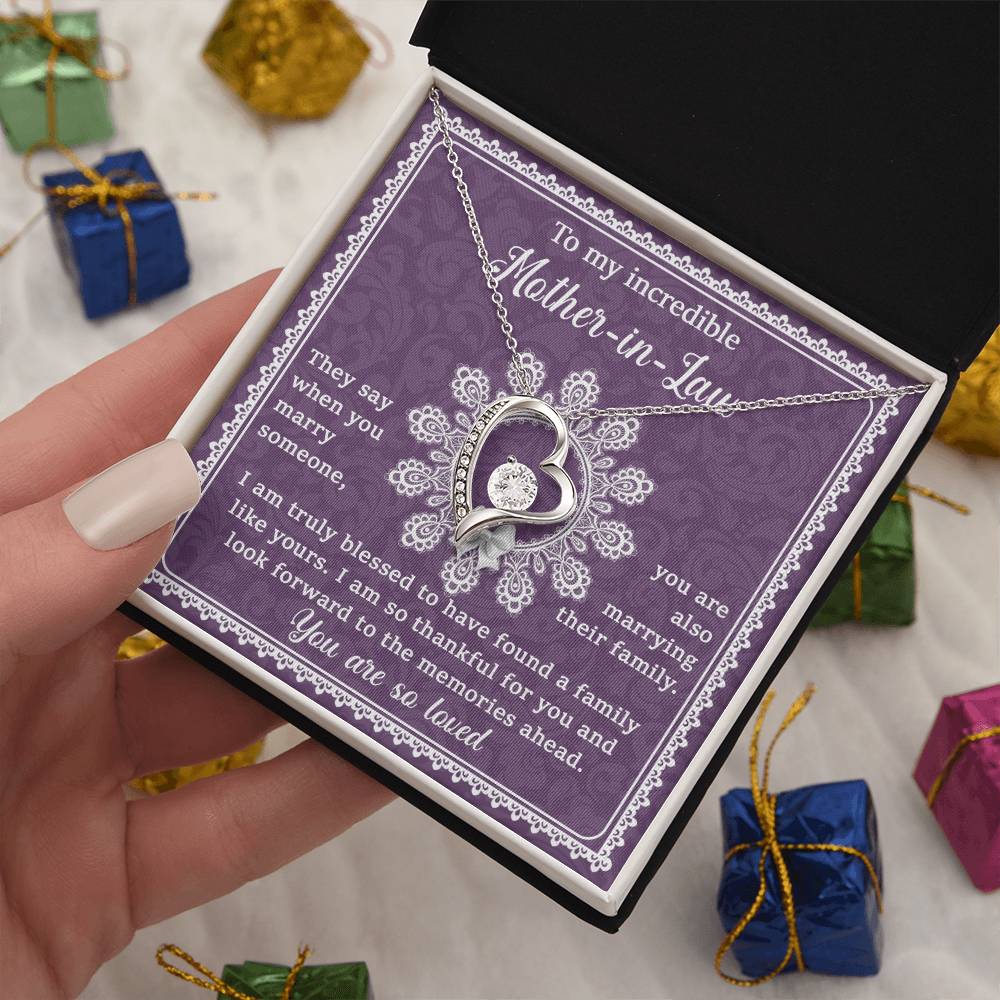 A person's hand holds a purple gift box containing the "To Mother-In-Law, Family Like Yours - Forever Love Necklace" with a 14k white gold finish and a heartfelt message card expressing appreciation for a mother-in-law. Small wrapped presents are visible in the background.