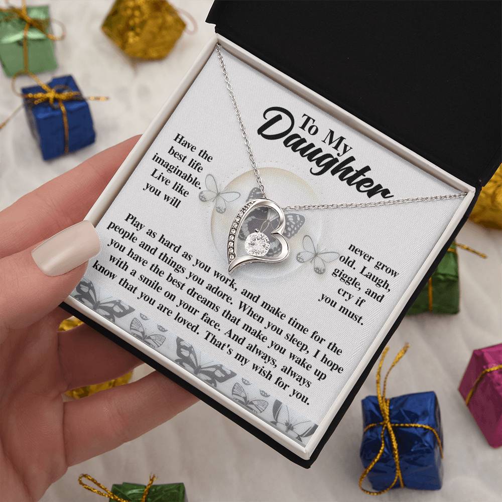 A close-up of the Daughter-My Wish - Forever Love Necklace is showcased with a gold finish, nestled in a gift box and complemented by a message card for a daughter. The necklace shimmers with cubic zirconia, while small wrapped presents enhance the backdrop's charm.