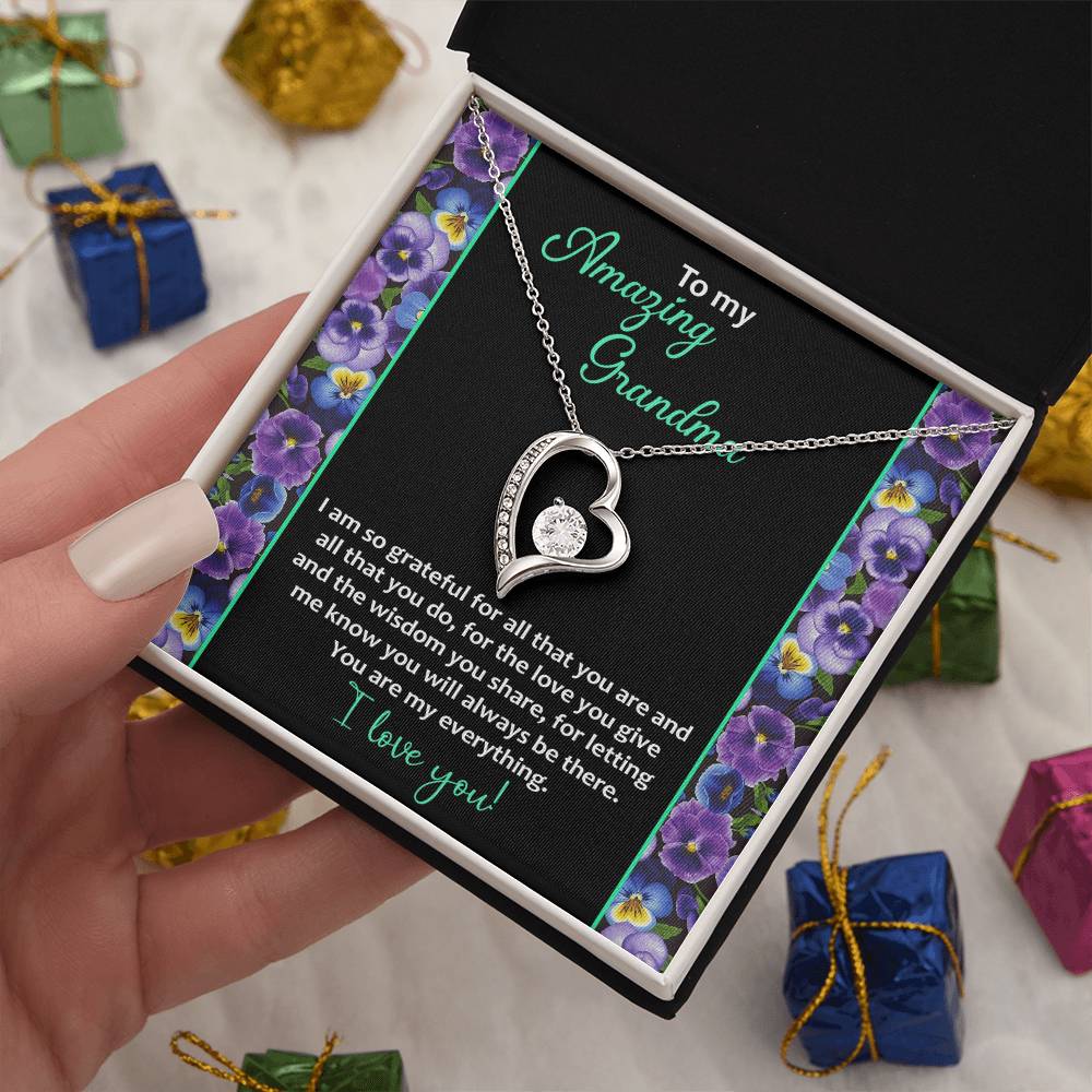 A hand holds an open jewelry gift box containing the To Grandma, My Everything - Forever Love Necklace, featuring a heart-shaped pendant in 14k white gold finish adorned with cubic zirconia stones. The card inside includes a heartfelt message to a grandmother. Small wrapped gifts are scattered in the background.