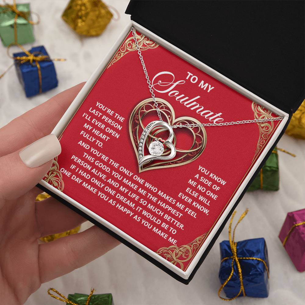 A hand holds an open gift box, unveiling the Soulmate-The Only One - Forever Love Necklace. This heart-shaped gem features a radiant gold finish on a red card with a heartfelt message. In the background, small wrapped gifts enhance this sentimental scene.