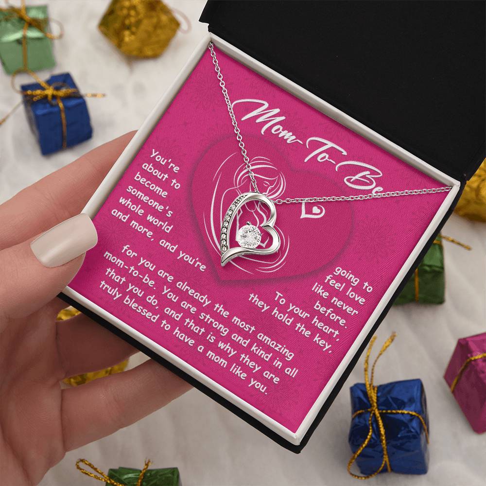 A hand holds an open box with a heart-shaped "To Mom To Be, Someone's Whole World - Forever Love Necklace" and a card that reads "Mom To Be," featuring a loving message for the expecting mother. The background showcases several wrapped Personalized Gifts.