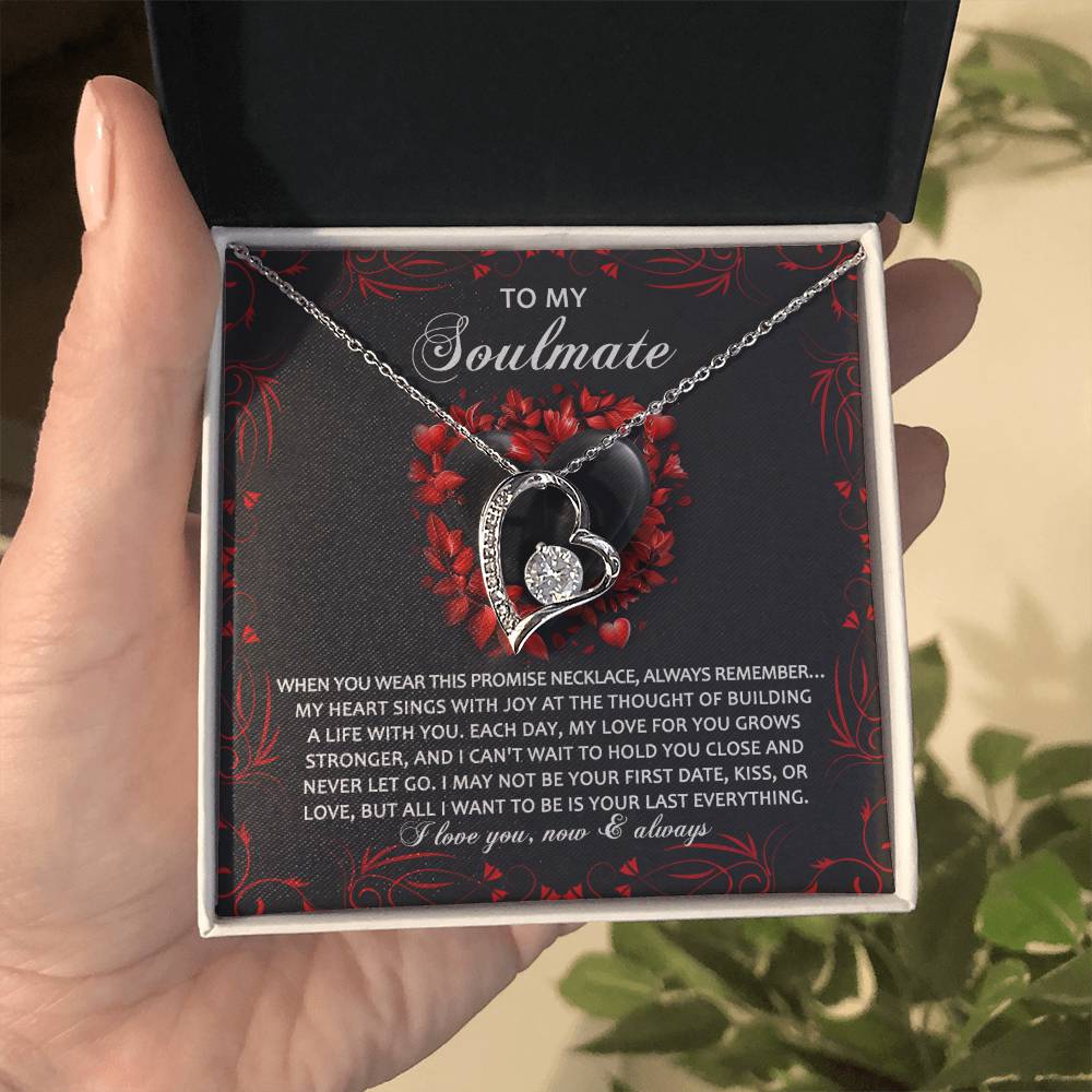 A hand opens a gift box unveiling the Soulmate-Hold You Close - Forever Love Necklace with a gold finish, a silver heart-shaped pendant, and sparkling cubic zirconia. The interior, adorned with red floral patterns, reads "To My Soulmate.