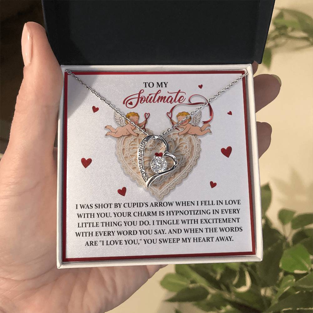 A hand holds an open jewelry box containing the Soulmate-Sweep My Heart - Forever Love Necklace, a heart pendant with a gold finish. The box displays a romantic love message, adorned with small hearts, cherub images, and sparkling CZ crystal accents.
