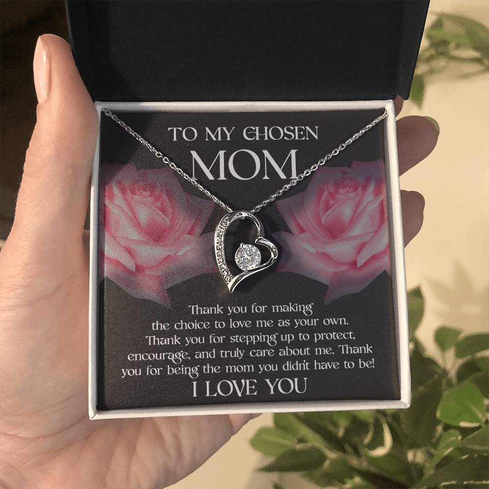 A hand holds a necklace box containing the "To Bonus Mom, My Chosen Mom - Forever Love Necklace," which features a heart-shaped pendant. The box reads, "TO MY CHOSEN MOM" with a heartfelt message and "I LOVE YOU" at the bottom. Available in white gold or yellow gold finish, it’s the perfect Mother's Day gift. Two pink roses are in the background.
