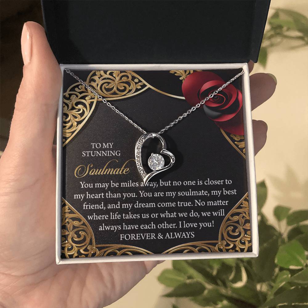 The Soulmate-Have Each Other Forever Love Necklace features a heart-shaped pendant with a central diamond, presented on a decorative card and accompanied by a romantic message. Its gold finish stands out against dark hues and red roses, creating an alluring backdrop.