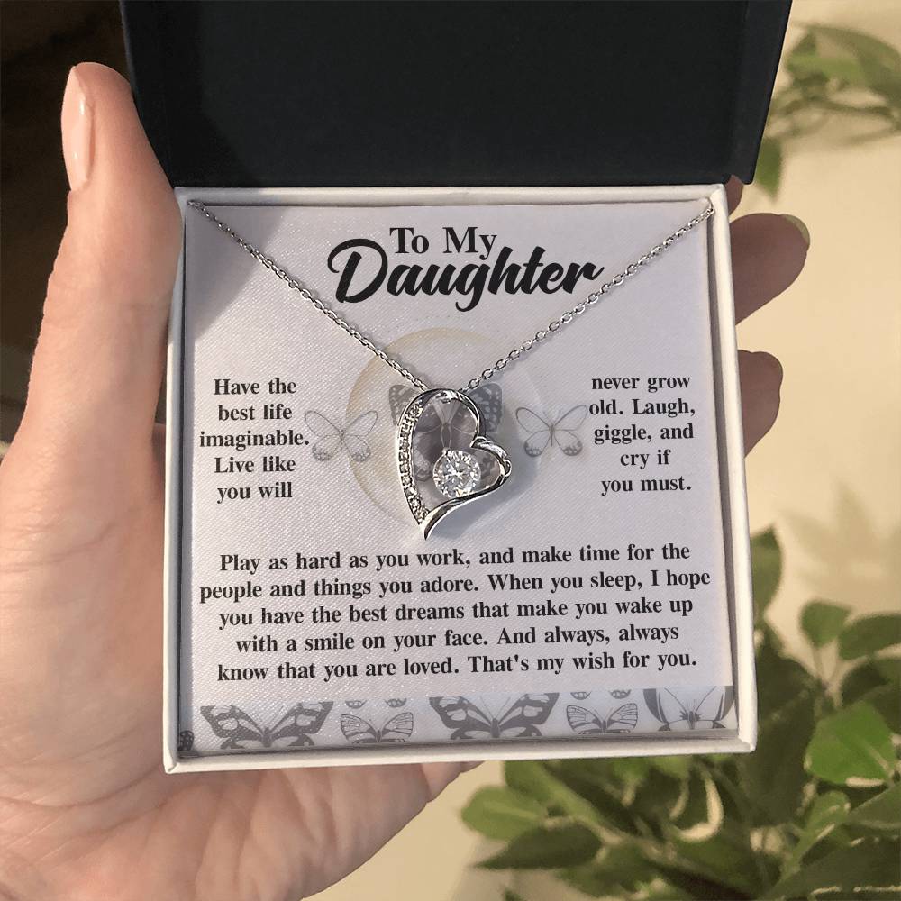 A hand holds a box displaying the Daughter-My Wish - Forever Love Necklace, featuring two interlocked hearts embellished with cubic zirconia. The gold finish shines elegantly, and the box lid includes an inspirational message dedicated to a daughter.