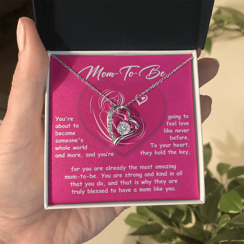 A hand holds a jewelry box containing the "To Mom To Be, Someone's Whole World - Forever Love Necklace" with a heart-shaped pendant, accompanied by a note that reads, "Mom-To-Be," expressing love and admiration for an expectant mother.