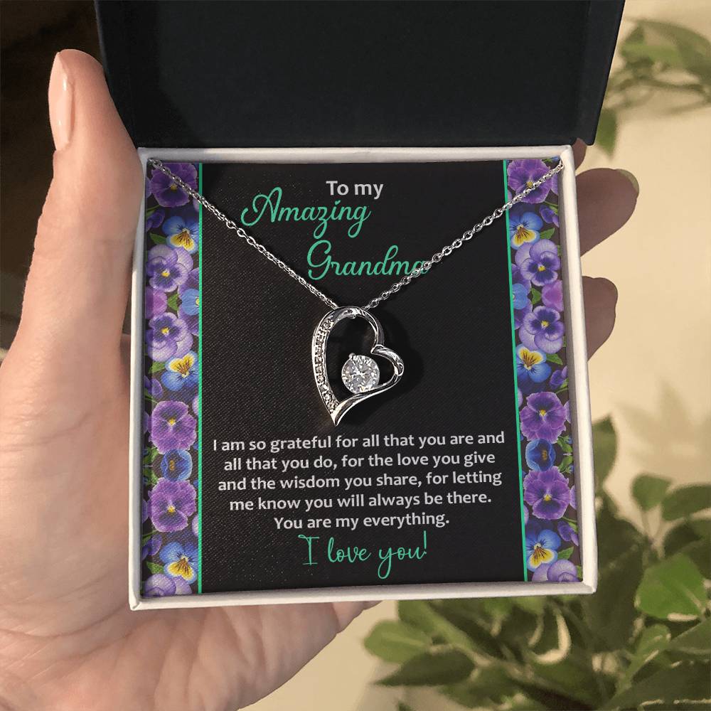 A hand holds the "To Grandma, My Everything - Forever Love Necklace" with a heart-shaped pendant inside a box that contains a message for "Amazing Grandma." Adorned in a 14k white gold finish and set with cubic zirconia stones, it rests against a backdrop of purple flowers.